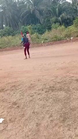 The video shows a person standing in a dirt road holding what appears to be a toy rifle and waving their arms, with another person standing further away in the background. The person in the foreground is wearing a hat, dark shirt, maroon pants, and sandals. The second person is wearing a light colored outfit and red hat or wrap. They are standing in a dirt road or open area surrounded by tropical vegetation and trees. The overall impression is of an outdoor scene with a casual or possibly playful interaction.