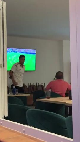 In a brightly lit indoor setting, possibly a restaurant or bar, two men are visible. The man on the left, wearing a white football jersey with Adidas stripes and black shorts, is using forearm crutches. He appears to be walking. He has dark hair and is looking towards the right.

On the right, a man wearing a pink polo shirt and glasses is seated at a table, facing away from the camera. He is looking towards a large television screen mounted on the wall. The television displays a football match, with players in red and white jerseys on a green field. Text overlays on the screen indicate a score and other game information.

Several tables with green upholstered chairs are arranged in the foreground, some with drinks and bottles on them. In the background, a row of empty chairs is stacked against the wall. The overall impression is of a casual viewing of a sporting event in a public establishment. The lighting suggests it is daytime, and the indoor setting implies no particular weather conditions are relevant. The man on crutches' presence might suggest an injury.