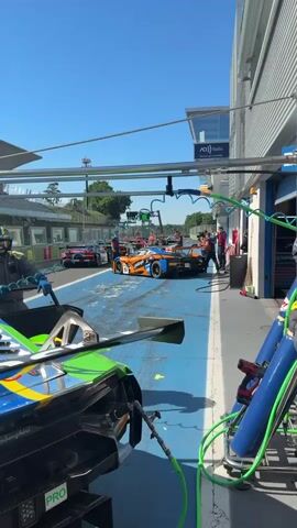 The video shows a race car in the pit lane of a race track. The car is orange and blue, with the number 59 on the side. Several people surround the car, presumably pit crew members, some wearing headsets. Other cars are visible in the background in adjacent pit areas. The pit lane is marked with blue paint, and there are overhead structures with lights and support equipment. The sky is clear and blue. A camera operator is filming the scene.