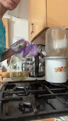 A person in a teal shirt is in a kitchen, standing in front of a gas stove. On the stove, there is a white enamel pot with a floral design. Above the pot, a large glass jar is being held by the person, tilted downwards as if to pour its contents into the pot. The person is holding the jar with a patterned cloth, likely to protect their hands from heat. The jar appears to contain a white substance. Steam is visible rising from the pot, indicating it is hot. Other kitchen items, including another glass jar and a rolling pin, are visible in the background. The kitchen has wooden cabinets.