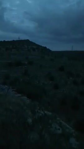 The video shows a dark, hilly landscape under a cloudy sky. The terrain is covered with sparse vegetation and rocks. In the distance, there are structures that appear to be power lines. The lighting suggests it is either dawn or dusk. There are no people or animals visible.