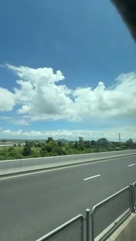 The video shows a view from a vehicle moving along a highway. The highway is paved with asphalt and has white lane markings. A metal guardrail is visible on the right side of the road. Beyond the highway, there is a landscape with green vegetation, trees, and fields. The sky is blue with white, fluffy clouds. The sun is shining, creating a bright atmosphere. A power pole and some distant buildings are also visible in the landscape. The video appears to be captured during the daytime.