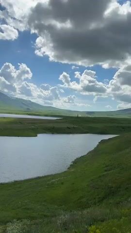 The video shows a serene landscape with a body of water, green hills, and a cloudy blue sky. The camera pans across the scene, capturing the reflections on the water and the vastness of the sky. The hills are covered in lush green grass, with patches of snow visible on the distant mountains. The clouds are dramatic, with some appearing dark and heavy, suggesting a possibility of rain, while others are bright and puffy. The overall atmosphere is peaceful and natural.