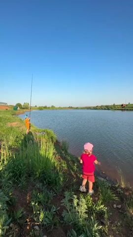 Two young children are at the edge of a small lake on a sunny day. A young boy in an orange shirt and a blue hat holds a fishing pole. He is turned toward the water. A young girl in a pink shirt and pink hat faces the water. She holds a small, translucent bucket. Tall green grass grows along the shoreline. In the background, across the lake, are a few small buildings. The sky is a clear, bright blue.