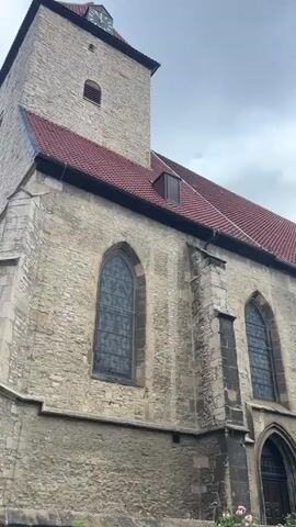 The video shows a low-angle exterior view of a stone church with a red tiled roof. A tall bell tower with a clock face is visible on the left side of the frame. The church has Gothic-style arched windows with stained glass. The sky is overcast, suggesting cloudy weather. The scene is static, with no visible movement or people.
