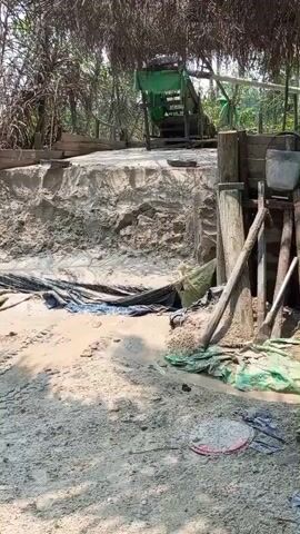 The video shows an outdoor scene, possibly a work area, with a sandy ground and some construction or equipment visible. In the background, there is a structure with a thatched roof, and a green conveyor belt system. To the right, there are wooden posts, a bucket, and some tools. There are also piles of sand and debris, including what appears to be a tire and some green tarp-like material. The overall impression is one of a rustic or informal operational setting.