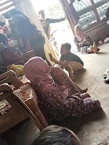 A group of people are gathered in a room. In the foreground, a woman wearing a pink hijab and a floral dress is sitting on the floor, leaning against a wooden chair. A young child in a dark t-shirt and light-colored pants is kneeling in front of her, holding a smartphone. Another woman in a pink hijab is sitting on the floor in the background, to the right. A man is standing near a doorway in the background, with his arms outstretched. The room appears to be a home, with wooden walls and furniture. Light is streaming in from the doorway, creating bright areas and shadows. The overall impression is of a casual gathering.
