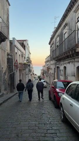 Three people walk down a narrow street paved with cobblestones. The street is lined with old buildings, some with balconies. Cars are parked along the side of the street. In the distance, the sea and sky are visible. The sky is a light pink and blue color, suggesting it is dawn or dusk.