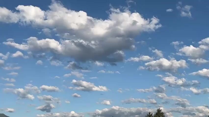 A video captures a time-lapse view of a bright blue sky filled with scattered white and grey clouds moving slowly across the frame. In the distance, the tops of lush green mountains and a few evergreen trees are visible, partially obscured by the clouds. The scene appears to be set during the daytime, with sunlight illuminating the clouds and the landscape below. The camera angle is fixed, looking upwards towards the sky, and the footage pans slightly to reveal more of the mountainous horizon. There are no people or specific objects that are the focus of the video, other than the dynamic movement of the clouds and the serene natural environment.