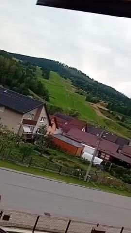 The video captures a tilted view of a residential area with houses and a street. In the background, a green, grassy hill slopes upwards, dotted with trees and a visible ski slope. The houses have tiled roofs and some feature window boxes with red flowers. A fence separates the grassy foreground from the street, and a few rocks are placed in the lawn. The sky is overcast with clouds.
