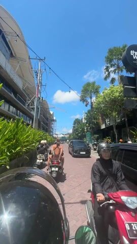 The video shows a first-person perspective from a person riding a moped down a brick-paved street in a tropical location. Another moped with two people, one of whom is wearing a swimsuit, is ahead. A black SUV passes on the right. The street is lined with shops, palm trees, and other tropical vegetation. A building under construction with a crane is on the left. The sky is blue with some clouds.
