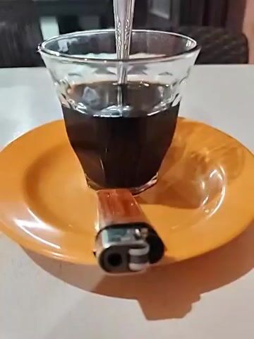 The video shows a glass of dark coffee with a spoon in it, sitting on an orange saucer. Next to the glass is a lighter. The table is white. In the background, there's a blurred view of a small outdoor restaurant or food stall, with food containers and various items on shelves. There are chairs visible as well.  The lighting is dim. The camera is static, with a slight zoom and focusing on the coffee cup and objects on the saucer.  There are some indistinct voices in the background.