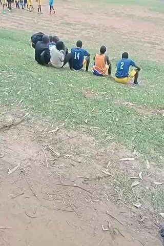 The video shows a grassy field with two groups of people. In the background, there is a group of people wearing soccer uniforms, some in red and some in blue and yellow. In the foreground, there is a group of six people sitting on the grass, watching the other group. One person is wearing a red shirt and has a black backpack next to them. Another person is wearing a blue shirt with the number 14 on the back. Another person is wearing a yellow shirt with the number 2 on the back. The field is surrounded by trees.