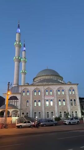 The video shows the front of a large mosque at dusk. The mosque is beige with gray accents and has two tall minarets that are lit with green and blue lights. There is a large dome on the main building. Cars are parked in front of the mosque, and a motorcycle is pulling away. The camera then pans to the right, showing a street with cars and a traffic light that is red. People are visible walking on the sidewalks. The sky is blue, and the lighting suggests it's either very early morning or late evening. The overall environment appears to be an urban or suburban setting in Turkey, indicated by the architectural style of the mosque.