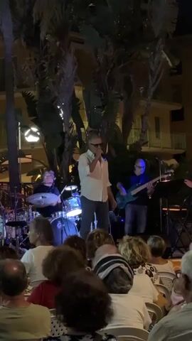 A group of people are watching a band perform outdoors at night. A male singer in a white shirt and sunglasses stands center stage, holding a microphone. To his left, a drummer plays a drum set. To his right, a man plays a bass guitar. The audience is seated in chairs facing the stage, with palm trees and a building visible in the background. The lighting is dim, with stage lights illuminating the performers. The video is shaky, suggesting it was filmed by someone in the audience.