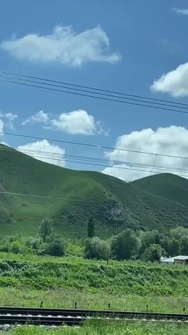 The camera is moving on a train, looking out of the window at a rural landscape. The sky is bright blue with white, puffy clouds. In the distance are green hills. There are several electric power poles and wires in the foreground. There are also trees and a few small buildings near the train tracks. A billboard with a child's picture on it appears briefly in the shot.