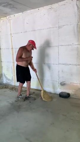 A shirtless man wearing a red baseball cap and black shorts is sweeping a concrete floor with a broom. The man is standing in what appears to be an unfinished or under-construction room, with bare concrete walls and ceiling. Debris is scattered on the floor around him, which he is gathering with the broom. 

The walls are constructed of large concrete blocks, and a yellow line is visible running vertically along the left wall, possibly indicating a utility line or marking. A dark, rectangular object rests against the right wall. The lighting suggests it is daytime, likely indoors, with light coming from an unseen source. The man's posture and the action of sweeping indicate he is engaged in cleaning the area. The overall impression is of a task being performed in a raw, undeveloped space.
