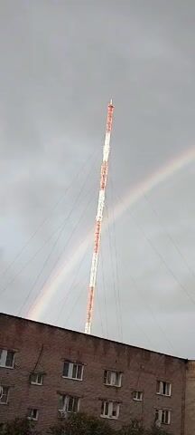 A cell phone tower with white and red sections stands tall against a cloudy sky. A faint rainbow arcs across the sky, partially obscured by the tower and a building's roof in the foreground. The building appears to be made of brick. The camera pans slightly to the right, revealing more of the rainbow and the cloudy sky, with the top of the cell phone tower still visible. Further to the right, a tree with dark green leaves and sparse branches comes into view. The camera continues to pan right, showing a longer portion of the rainbow and the overcast sky. The bottom of the frame shows the tops of more buildings and some leafy branches of a tree.