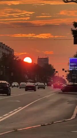 A sunset is viewed over a city street with traffic. The sun is a bright orange orb partially obscured by clouds and buildings. The sky is a vibrant orange and yellow, with scattered clouds. Cars are driving down a multi-lane road, with streetlights and buildings visible on either side. The overall mood is peaceful and serene.