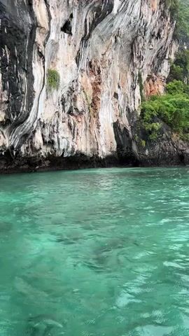 The video shows a boat moving through clear turquoise water near a large, sheer rock face. The rock face is weathered and layered, with patches of greenery growing on it and at its base. The water is calm and shows some reflections of the rock and sky. The overall scene is of a natural, tropical landscape.