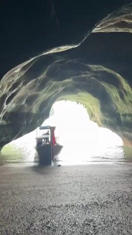 The video shows a view from inside a dark cave looking out towards a brightly lit body of water. A small, traditional wooden boat with a red canopy is partially submerged in the water near the cave entrance. The cave walls are rough and uneven. The water is calm and appears greenish, with distant limestone karsts visible in the background. The ground in the foreground is wet and pebbly. The camera is stationary.