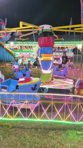 The video shows an amusement park ride with various colorful, cartoonish animal-shaped seats, including a blue cat, a white and blue zebra, and a blue horse. The ride is illuminated with neon lights in green and pink, creating a festive atmosphere. In the background, a larger blue inflatable slide is visible, along with several people sitting and standing, and a child walking. Several other rides and game stalls are also present in the background, indicating a bustling fairground. The ride appears to be in operation, with the central column rotating and the animal seats moving. The ground is covered in grass, and the sky is dark, suggesting it is nighttime.