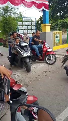 Two scooters are parked at an entrance of a building. The building has a blue facade with red and white decorations and is labeled "SOY KREUEN." A man is riding a dark gray scooter, carrying two young children. Another man is riding a red scooter, carrying one child. Several other people are on motorcycles in the background, some appearing to be students. The scene suggests a school drop-off or pick-up.