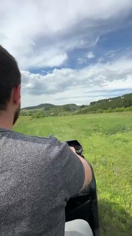 A man wearing a gray patterned shirt is riding a motorcycle across a grassy field on a partly cloudy day. Mountains are visible in the distance. The camera's perspective is behind the driver, showing his head and torso, as well as the motorcycle's rearview mirror. The camera shakes as the motorcycle moves.