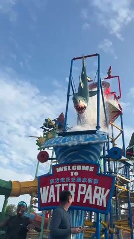 The video shows a scene at a water park. In the foreground, two men are visible. One man on the left is wearing a black tank top and sunglasses, and he is smiling and looking upwards. The other man on the right is wearing a gray long-sleeved shirt and is also looking upwards. Behind them, a large water slide structure is prominent. A shark figure is suspended upside down at the top of the structure, with water cascading from it. A sign that reads "WELCOME TO WATER PARK" in red and white letters is attached to the structure. The sky is blue with some clouds. Various parts of the water slide, including colorful tubes and metal supports, are visible. Water is spraying from different points on the structure, creating a dynamic and wet environment.