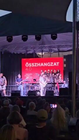 A band performs on a stage under a canopy. A large red screen behind them displays "ÖSSZHANGZAT 4.0" in white text, with a group photo of band members below. Two men are on the left side of the stage, one singing into a microphone and playing a guitar, and the other standing by a keyboard. A man in the center plays a saxophone. Other band members are visible behind him and to the right, some holding instruments. Drums and other musical equipment are on the stage. An audience is seated in front of the stage, facing the performers. The lighting is stage lighting, with some colored lights visible.