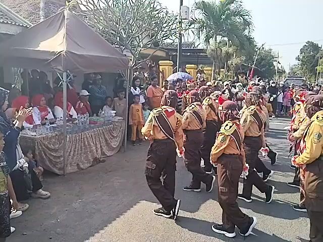 A group of scouts in brown and tan uniforms are marching in formation on a paved street. They are wearing maroon berets and have tan sashes. There is a tent set up on the left side of the street with people sitting under it, some of whom appear to be judges. A crowd of spectators lines the street. There are trees and buildings in the background. The sky is visible and appears to be daytime with bright sunlight.