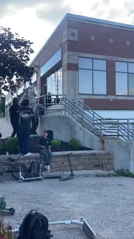 The video shows a group of young people gathered outside a brick building. Some are on scooters and BMX bikes. One person is attempting a trick down a set of stairs on a scooter. Others are watching and filming. The environment appears to be an urban or suburban outdoor space with a parking lot and some greenery. The weather is cloudy.