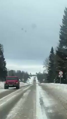 The video shows a car driving on a snow-lined road. A red Jeep Wrangler passes by. There are trees on either side of the road, and signs are visible. The sky is overcast and gray. The road is wet and icy. The video is taken from inside a car, looking forward. The road slopes upward in the distance. There are traffic lights at the top of the hill.