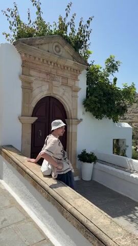 A person sits on a low white wall next to a white building with an ornate door. The view pans from the person to a bay filled with people, boats, and a beach. The landscape includes a rocky, grassy hillside. The sky is clear and blue.