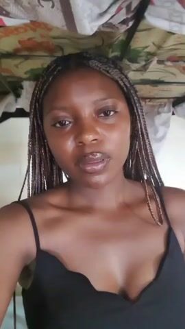 A young woman with braided hair, wearing a black tank top, looks at the camera.  Her skin appears shiny. The background shows a white sheet with a colorful floral pattern. The camera remains static.