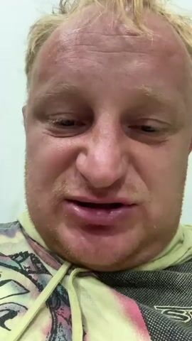 The video is a close-up of a light-skinned man with blond hair and a beard. He is wearing a hooded sweatshirt. The man appears to be speaking to the camera, though his mouth is partially obscured. His expression is difficult to discern due to the low resolution and close cropping of the video. The background is a plain, light-colored wall.