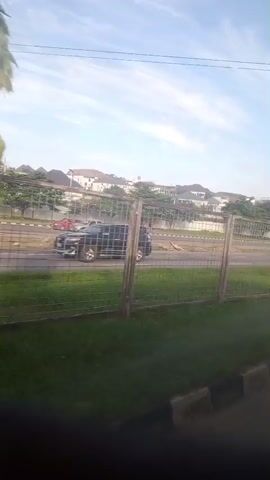 The video shows a view from inside a vehicle looking out at a road and some buildings in the background. The camera pans slightly to the right. On the right side of the road, there is a fence and some trees. Several cars and a bus are visible on the road. The sky is blue with some clouds.