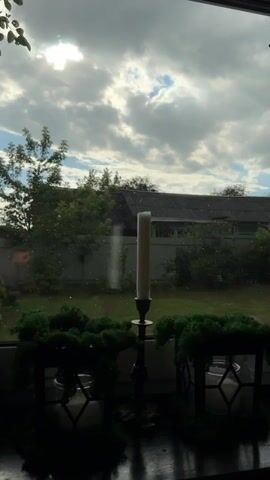 This media file depicts a scene viewed through a window, looking out onto a backyard. The foreground is dark and reflective, showing the silhouette of a table with decorative items. A tall, unlit white candle in a black candlestick stands prominently in the center of the view. To the left and right of the candlestick are dark planters filled with vibrant green moss.

Beyond the window, a garden setting is visible under a dramatic sky. Lush green trees and bushes form a natural backdrop, with a white fence running horizontally across the mid-ground. A dark-colored roof of a building or shed is prominent above the fence. The sky is filled with large, fluffy white clouds interspersed with patches of blue, and the sun is partially obscured by the clouds, suggesting it might be late afternoon or a partly cloudy day. A few small white specks are visible on the windowpane, possibly dust or rain.

The overall mood is tranquil and domestic, with a touch of natural beauty from the garden and sky. The unlit candle suggests a quiet moment, perhaps before an evening gathering or simply a decorative element. The location context provided places the scene in Petrovskoye, Russia. There is no visible text in the media file.