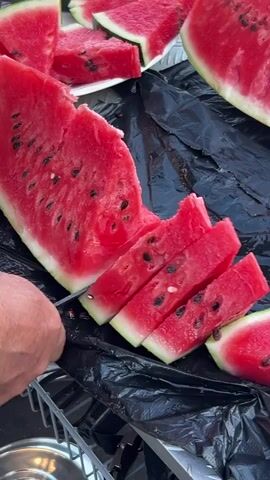 A close-up shot shows a person's hand using a black-handled knife to cut slices of watermelon. The watermelon slices are arranged on a black plastic sheet. Several other watermelon slices, some on a white plate, are visible in the background. The scene appears to be outdoors, possibly at a picnic or market, given the presence of a metal rack and what looks like a sink in the lower left corner. The watermelon is ripe, with visible seeds.