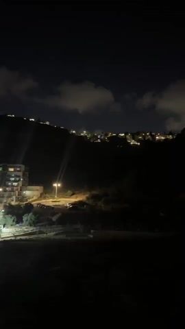 A nighttime view shows a dark, hilly landscape with lights from a distant town illuminating the horizon. Clouds drift across the night sky, partially obscuring the stars. In the foreground, a street lamp casts a bright beam onto a dirt road, illuminating a small building. The overall scene is dimly lit, creating a peaceful and serene atmosphere.