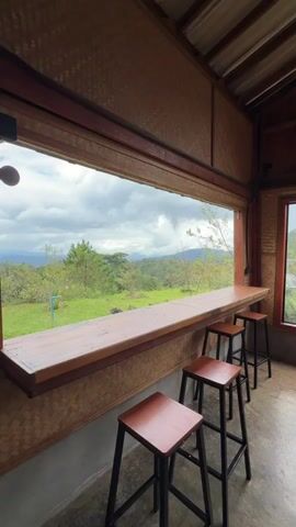 A video pans across a rustic indoor seating area with a large window offering a panoramic view of a green, hilly landscape under a cloudy sky. The foreground features a long, polished wooden counter with three dark-seated, black-legged bar stools positioned in front of it. The walls are covered in a woven, natural material, possibly bamboo or straw, and the ceiling appears to be corrugated metal. The window is wide and low, creating a seamless transition between the indoor space and the outdoor scenery. The view outside includes rolling hills, scattered trees, and a dense forest in the distance, all bathed in natural, diffused light.
