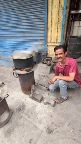 The video shows a man in a pink shirt and jeans squatting beside a large pot of boiling liquid on a charcoal stove. He is holding a metal rod and appears to be tending to the fire. Another person walks into the frame wearing a white shirt and pants. The scene appears to be outdoors, possibly on a street, with a blue shuttered building in the background. There are other metal objects and debris on the ground, suggesting a work or street environment.