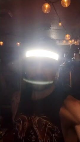 Here's a description of the video:

The video features a man in a dimly lit room, possibly a workshop or garage. He is wearing a black tank top with a graphic design and has visible tattoos on his arms. A bright LED headband is positioned on his forehead, obscuring much of his face due to the intense light it emits. He has a mustache and appears to be speaking or making expressions. In the background, several decorative light bulbs hang from the ceiling, contributing to the overall mood of the scene. There are also various items and equipment visible, suggesting a workspace or storage area. The video is shot from a low angle, looking up at the man.