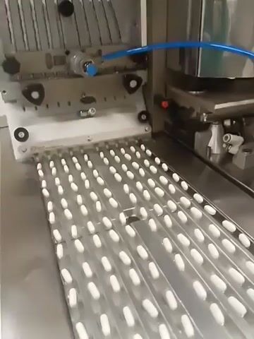 A metal, industrial machine fills blister packs with white, oval pills. The packs are silver and lined with multiple rows, each containing several pills. A mechanical arm with multiple prongs pushes the pills into the blisters. A blue tube is connected to the arm, likely providing pneumatic power. The machine is in a clean, industrial setting.