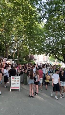 The video shows a busy outdoor scene, likely a park or public square, filled with people. Tall, leafy trees provide shade. In the foreground, a paved path runs alongside a sandy area where many people are gathered at small tables, some eating or drinking, others socializing. A few people are walking on the path, while others are milling about in the background. The atmosphere appears relaxed and convivial. The lighting suggests it is daytime, possibly late afternoon or early evening, with the sun filtering through the trees. Some stalls or structures are visible in the far background, hinting at an event or market.