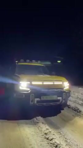 At night, a yellow electric pickup truck is stopped on a snowy dirt road, its headlights shining brightly. The truck has a light bar on its roof, and white running lights across the front grill.  The license plate reads "FE003 AK-4". The sound of the vehicle's horn is heard.