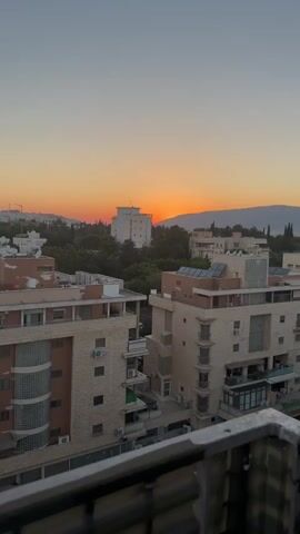 The video captures a sunset over a city with apartment buildings and trees. The sky displays a gradient of orange and yellow hues. The sun is a bright orange disc partially obscured by a mountain range in the distance. Several apartment buildings, some with balconies and air conditioning units visible, are in the foreground and middle ground. Lush green trees fill the spaces between the buildings. The scene is viewed from an elevated position, possibly a balcony, with a railing in the very bottom foreground. No people are visible in the video.