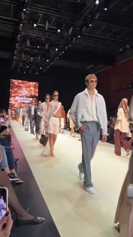 The video shows a fashion show with models walking down a runway. The runway is lit and there is a large screen at the back with the words FASHION SHOW. There are people in the audience watching the show. The models are wearing various outfits, and some of them are carrying bags. The scene appears to be indoors.