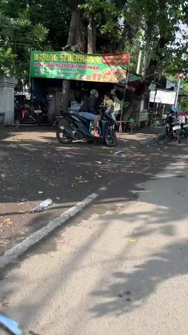 The video shows a street scene with a food stall and several motorcycles. A person on a black motorcycle is parked in front of the stall, facing the camera. The stall has a green sign with text in Indonesian, indicating it sells fried foods and drinks. There are other motorcycles parked around the stall, and trees provide shade. The ground is paved and appears somewhat worn. The lighting suggests it is daytime.