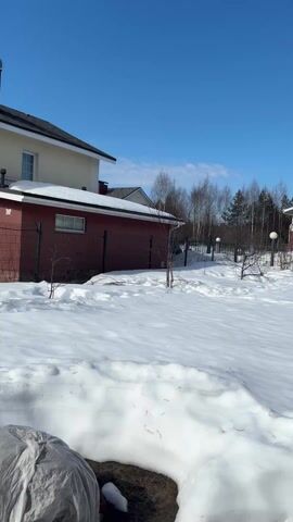 The video opens with a static shot of a snow-covered landscape under a clear, bright blue sky. In the immediate foreground, a large mound of snow is partially obscured by a grey, tarp-like covering. To the left, the side of a house with a dark red lower half and a cream-colored upper half, topped with a dark roof, is visible. A dark metal fence runs in front of it, behind which are some bare shrubs or small trees. In the background, a dense line of bare deciduous trees stands against the horizon.

At the 0:01 mark, the camera begins a slow pan to the right. The first house gradually moves out of frame. As the camera pans, a second, larger house comes into full view. This house shares a similar architectural style, featuring a dark red lower section and a light cream or yellow upper section. It boasts a prominent second-story balcony with a white railing, and a ground-floor porch or entrance area with a similar white railing and white-framed windows. A black metal fence separates the house from the wide, snow-covered lawn. Two white globe-shaped garden lamps are visible behind the fence. The background continues to show the bare trees and the unblemished blue sky.

Around the 0:03 mark, the pan continues, bringing the camera closer to the side of the first house that was partially seen at the beginning. This house now shows more detail: its lower wall is dark red, transitioning to a section of stone cladding, and then to a cream-colored upper wall. Two white-framed windows are visible on the red wall. To the right, there's an enclosed white porch or entrance with steps leading up to it, and a white-framed window above. A narrow strip of brown ground is visible along the house's foundation, where the snow has melted or been cleared. The video ends at 0:05, still focused on this section of the house, with the vast expanse of snow and clear blue sky dominating the frame. The overall impression is a sunny, late winter or early spring day in a residential area.